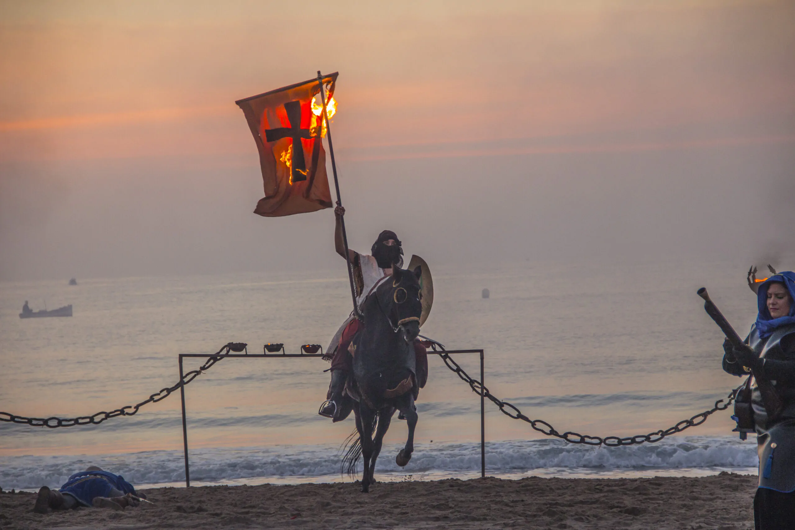 Caballero medieval montado a caballo ondeando una bandera en llamas al atardecer durante una recreación histórica en la playa