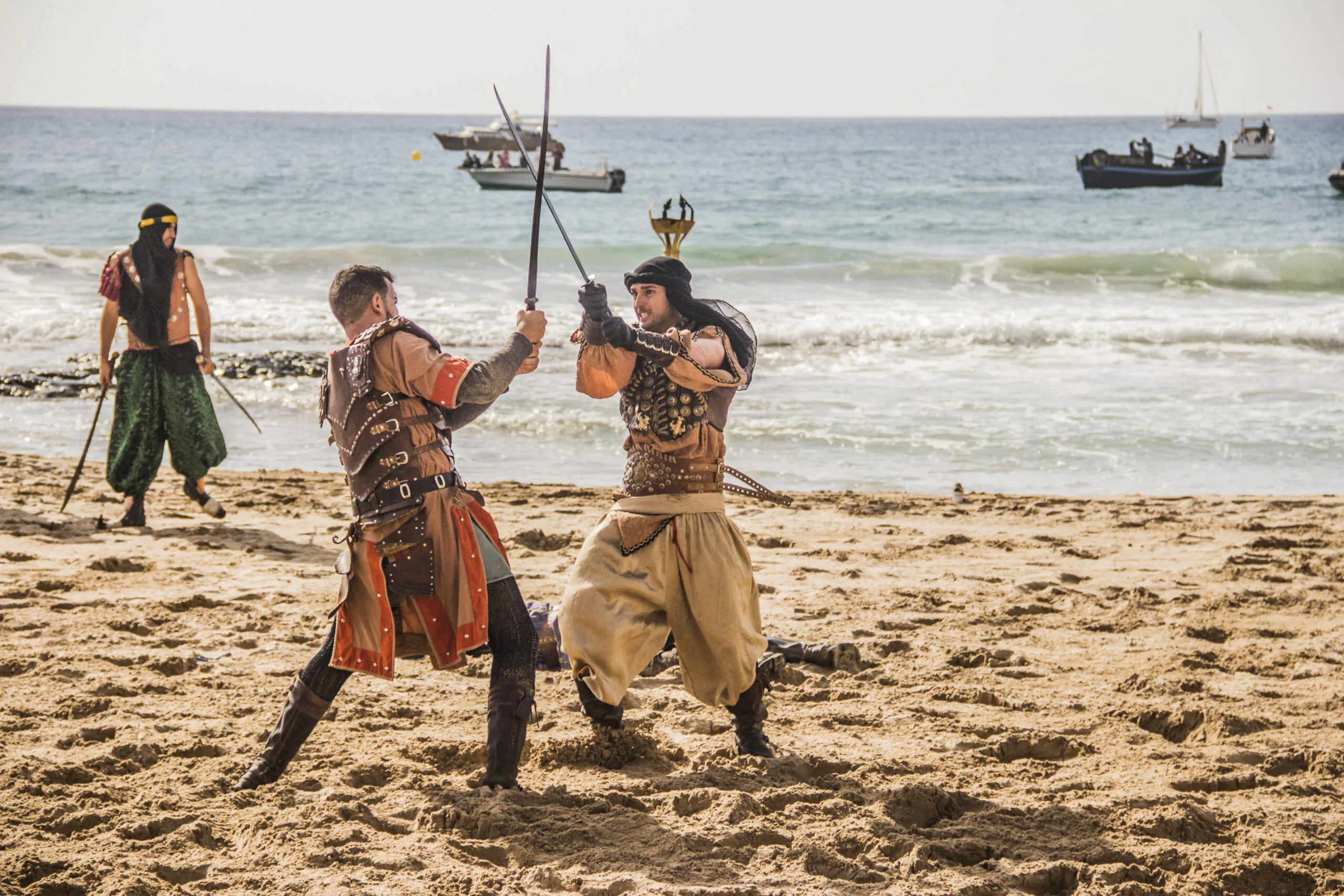 Guerreros medievales combatiendo con espadas en una playa, mientras otros observan y barcos navegan en el fondo