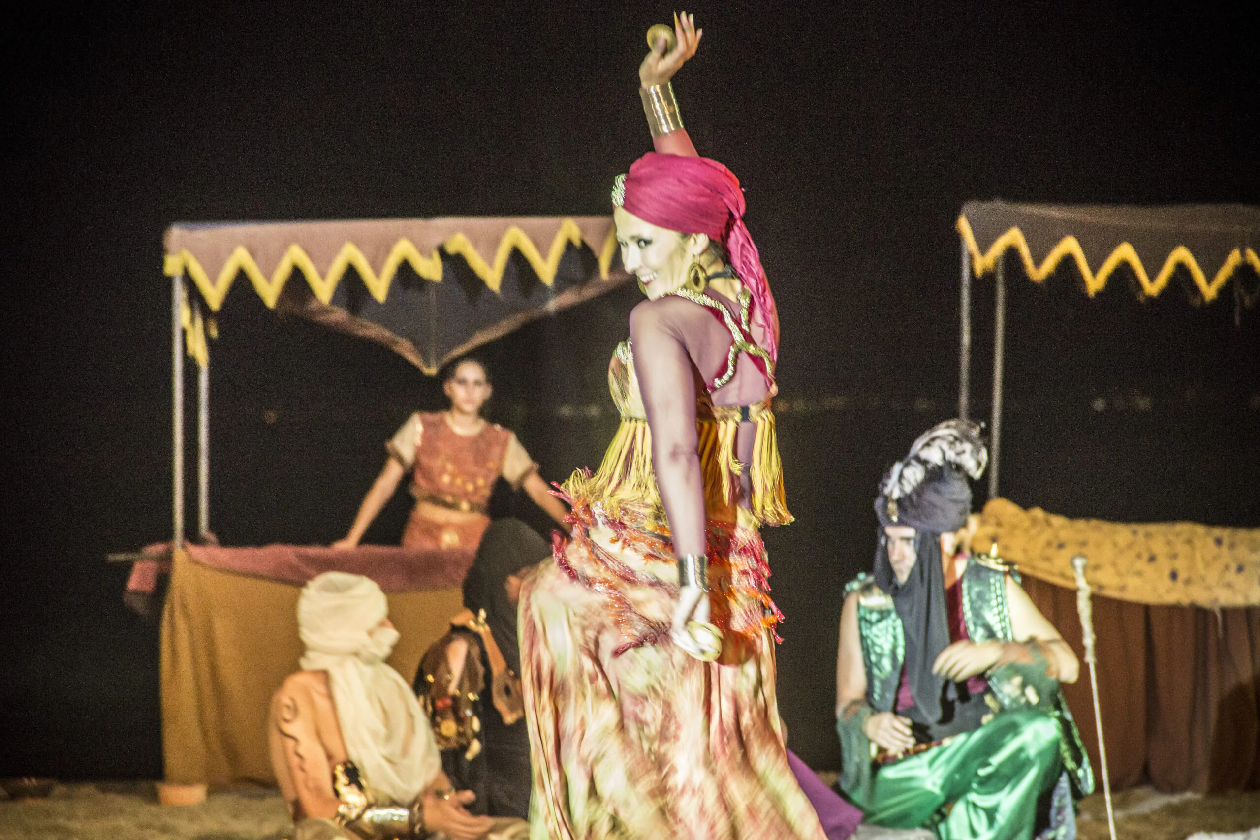 Bailarina con vestimenta tradicional actuando en un escenario al aire libre durante la noche, rodeada de personajes con trajes orientales