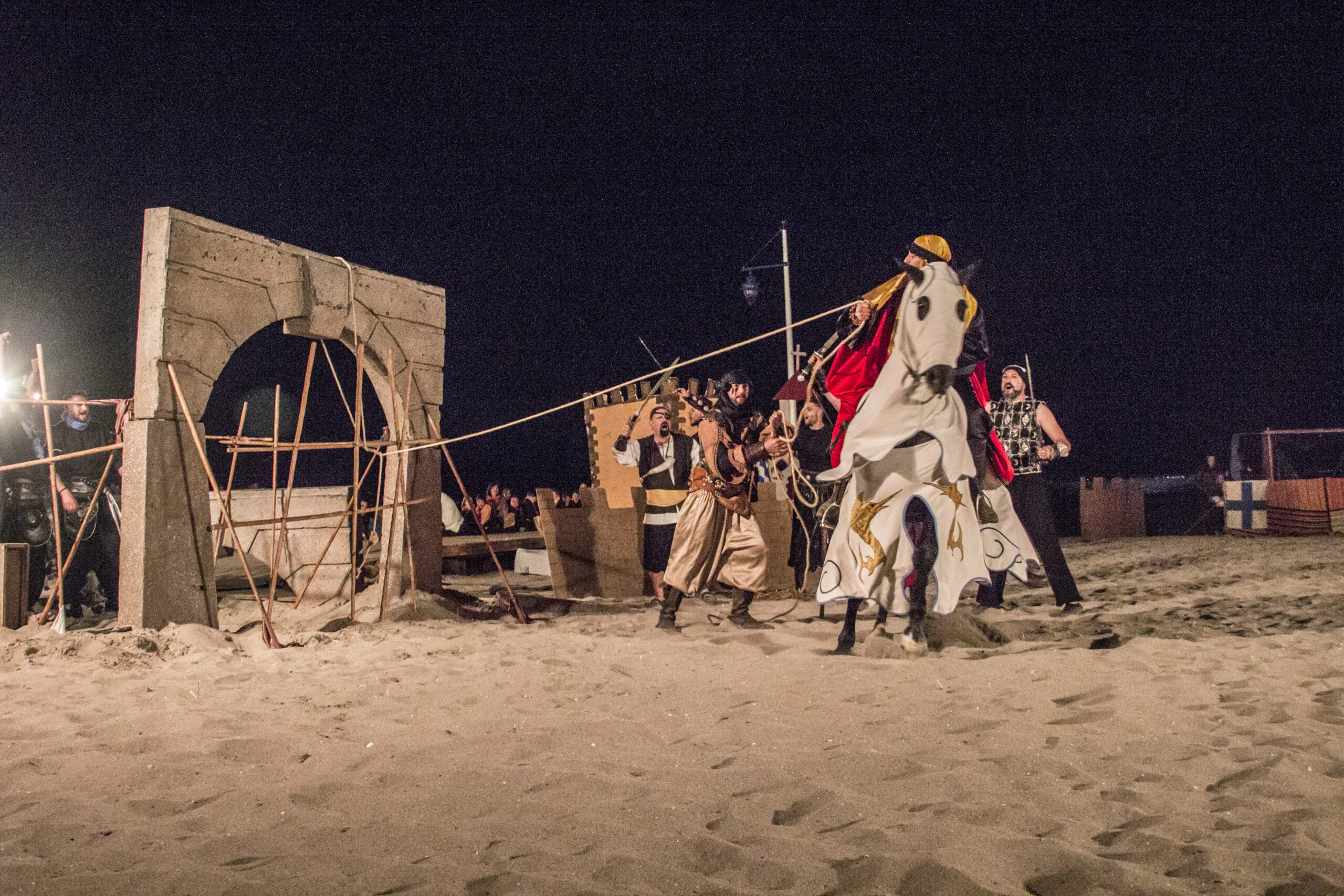 Recreación nocturna de una escena medieval en la playa, con un caballero a caballo y actores tirando de cuerdas para derribar una estructura