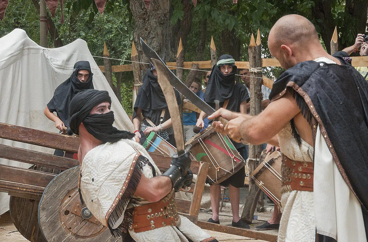 Recreación histórica de una batalla con combatientes vestidos con túnicas y espadas, acompañados por músicos tocando tambores en un campamento medieval