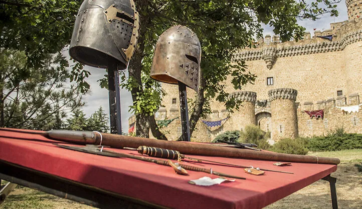 Exhibición de cascos medievales y armas antiguas sobre una mesa roja, con un castillo de fondo en un evento histórico al aire libre