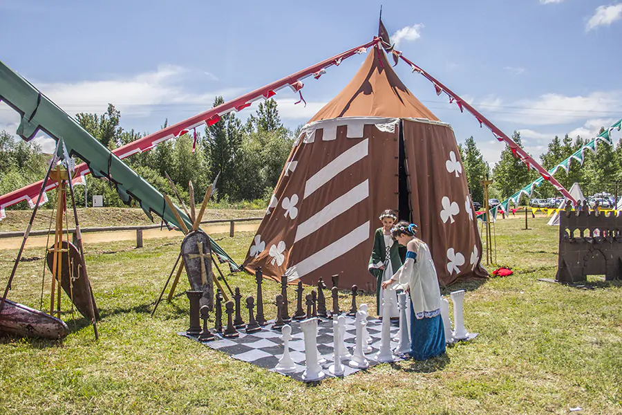 Dos niños vestidos con trajes medievales jugando ajedrez gigante frente a una tienda decorada en un campamento temático