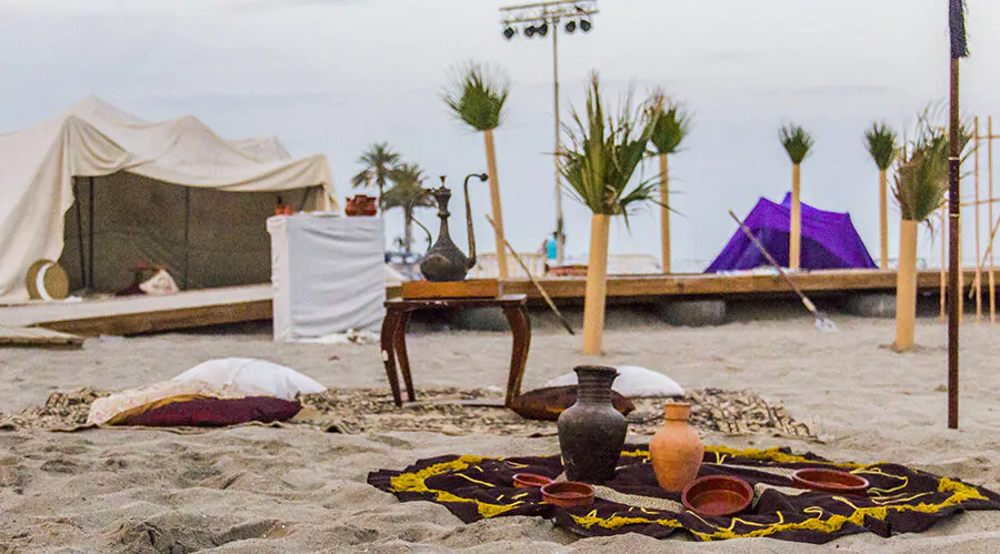 Escena de campamento histórico en la playa con alfombras, cojines y cerámica dispuestos en la arena. Al fondo, se aprecian tiendas de campaña de estilo antiguo, palmeras decorativas y un escenario de madera, con un cielo nublado que proporciona un ambiente tranquilo y atemporal