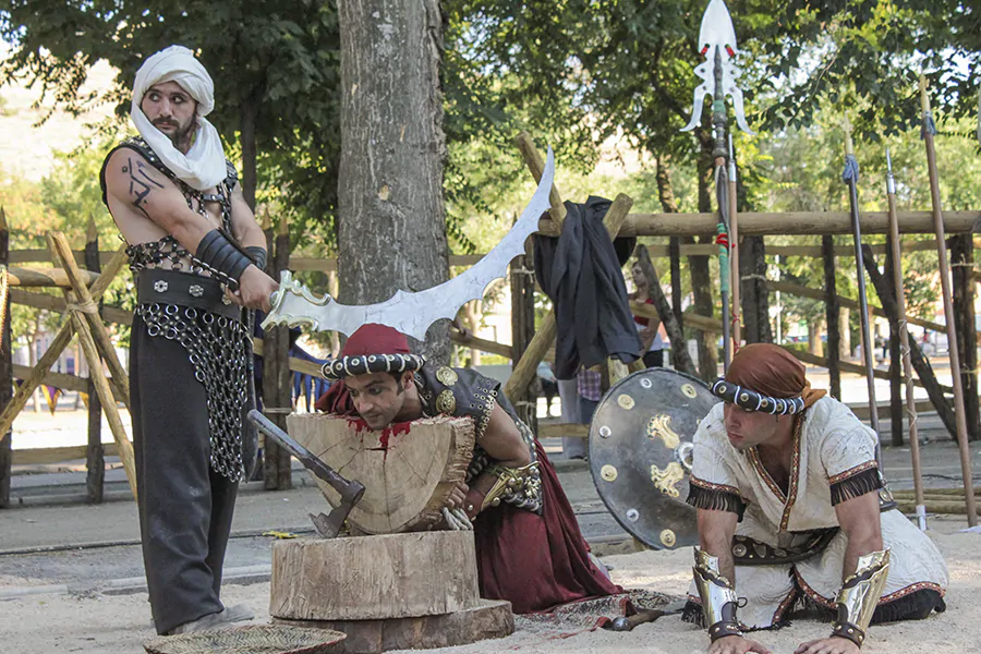 Recreación histórica con tres actores vestidos con trajes medievales, uno de ellos sosteniendo una gran espada curva mientras los otros dos se arrodillan frente a un tronco de madera