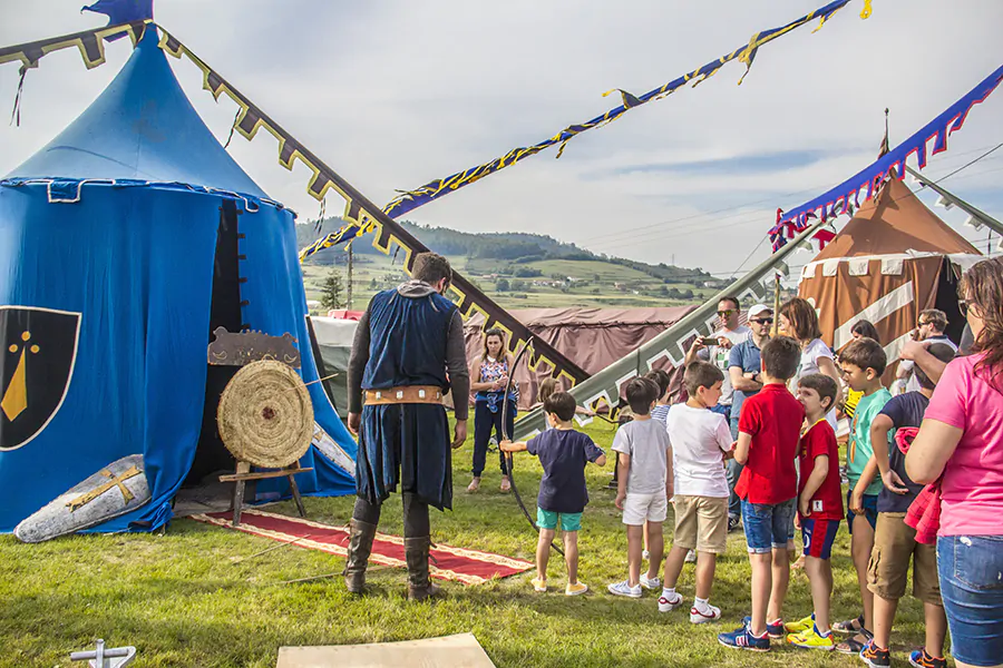 Niños reunidos en un campamento medieval participando en una actividad de tiro con arco, con tiendas y decoración temática de fondo