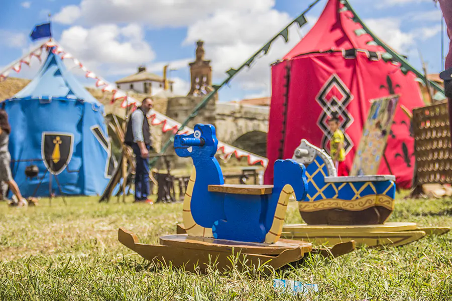 Caballitos de madera en forma de dragón y caballo en un área de juegos en un campamento medieval, con tiendas coloridas de fondo y decoración temática