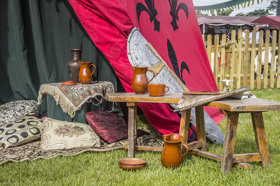 Escena de campamento medieval con jarras de barro, cojines decorativos y un escudo apoyado en una tienda roja adornada con lirios