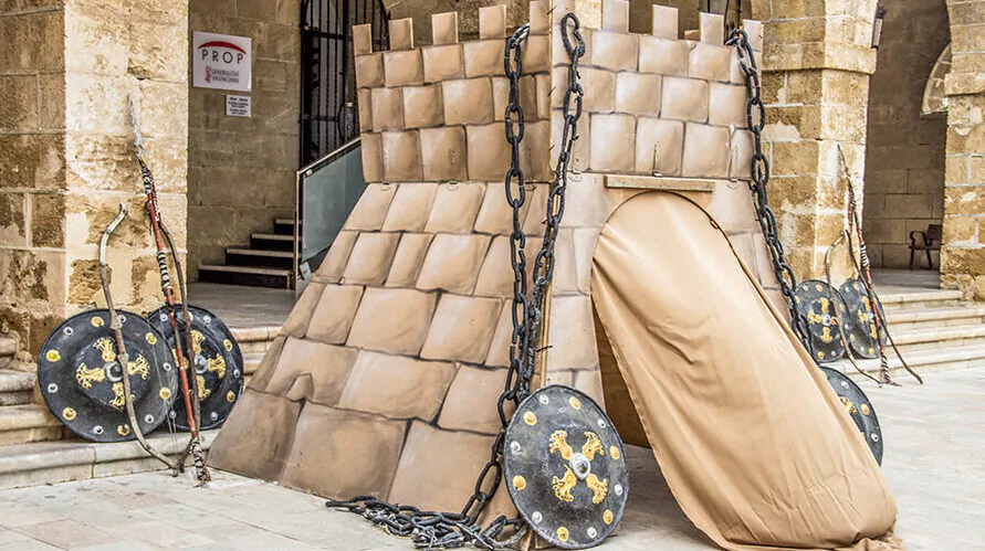 Recreación de una estructura medieval en forma de torre, decorada con cadenas y escudos con motivos dorados. La torre está ubicada en un entorno histórico con arcos de piedra y escaleras, lo que sugiere un evento de recreación o feria medieval