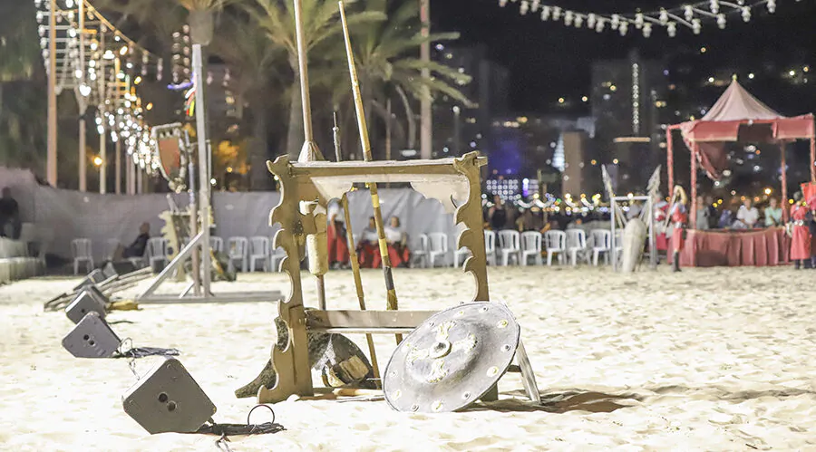 Escenario en la playa por la noche con equipo medieval, incluyendo un soporte de madera con armas y un escudo. En el fondo se ven palmeras, luces de la ciudad y carpas decorativas. La disposición sugiere una recreación histórica o un evento medieval al aire libre