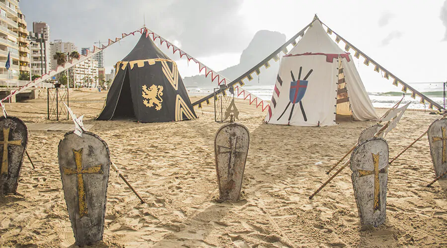 Campamento medieval recreado en la playa, con tiendas de campaña de estilo histórico decoradas con escudos y emblemas. En primer plano, varios escudos con cruces doradas están alineados en la arena. Al fondo, se observa el mar y un paisaje de edificios modernos, creando un contraste entre la recreación histórica y el entorno urbano