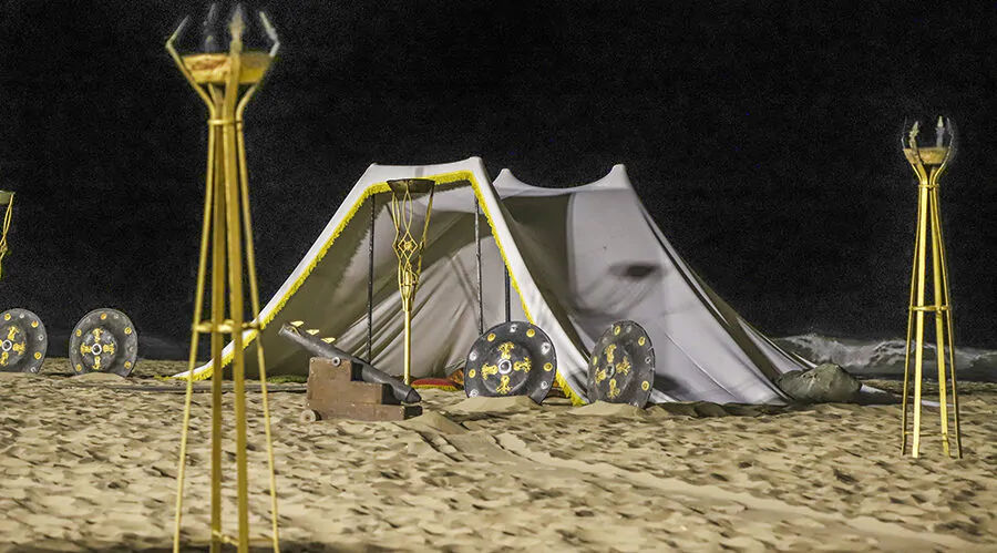 Campamento nocturno en la playa, con una tienda de campaña blanca decorada con bordes amarillos y rodeada de antorchas altas. Escudos decorativos con detalles amarillos y negros están dispuestos frente a la tienda, y el ambiente sugiere una ambientación medieval o histórica